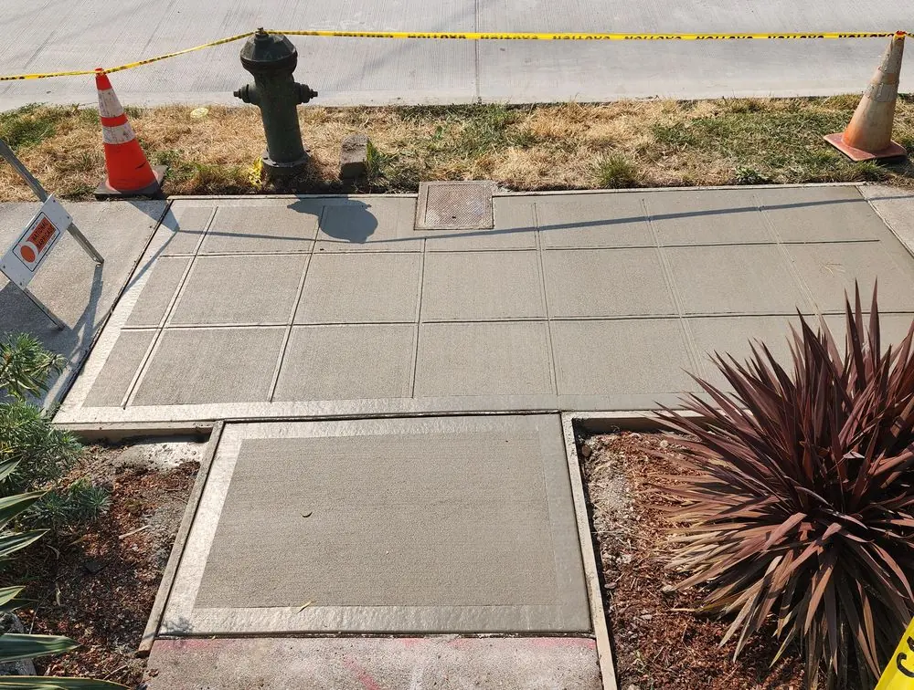 Finished sidewalk panels along a street, with traffic cones holding the work area and a work truck farther down the block.